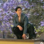Young man in stylish attire poses outdoors with vibrant jacaranda tree backdrop.