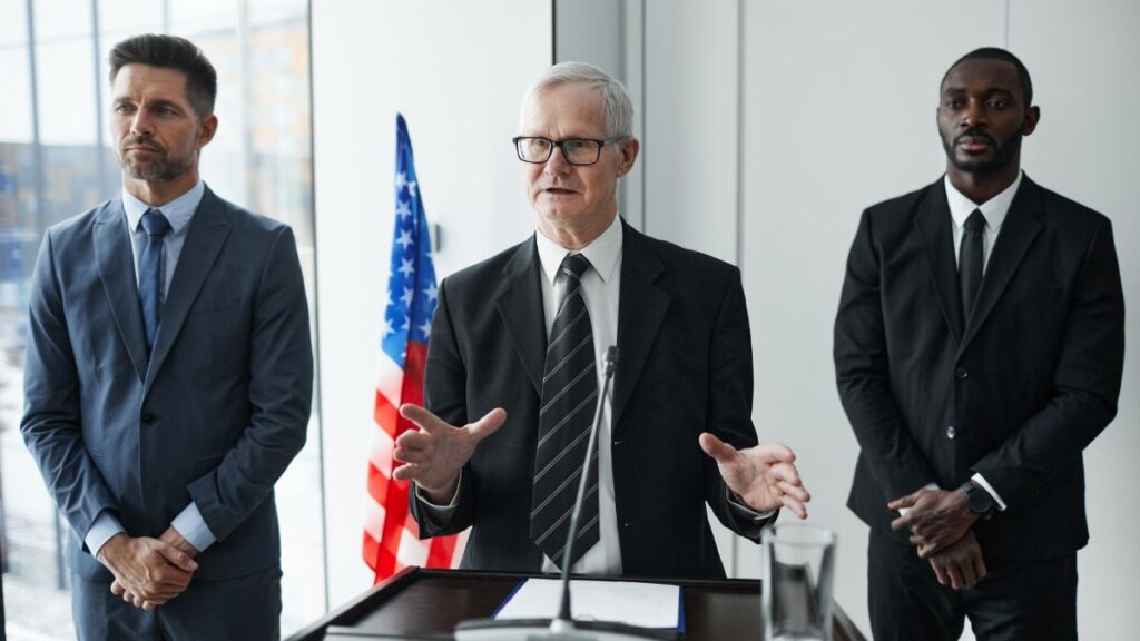 Three business professionals giving a speech with an American flag backdrop.