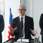 Three business professionals giving a speech with an American flag backdrop.