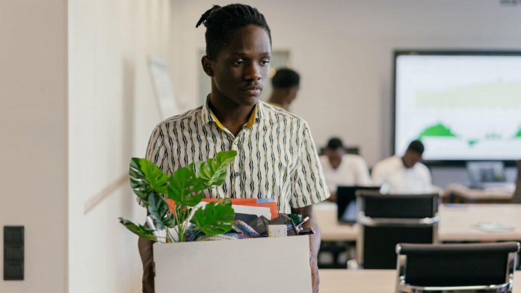 A serious young man carrying a box of personal items in an office setting, suggesting dismissal or resignation.