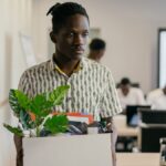 A serious young man carrying a box of personal items in an office setting, suggesting dismissal or resignation.