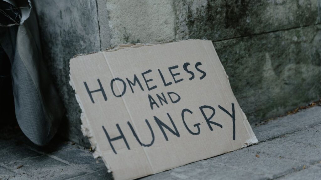 Cardboard sign with 'Homeless and Hungry' message leaning against a wall on a sidewalk.