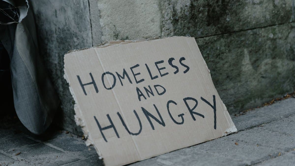 Cardboard sign with 'Homeless and Hungry' message leaning against a wall on a sidewalk.