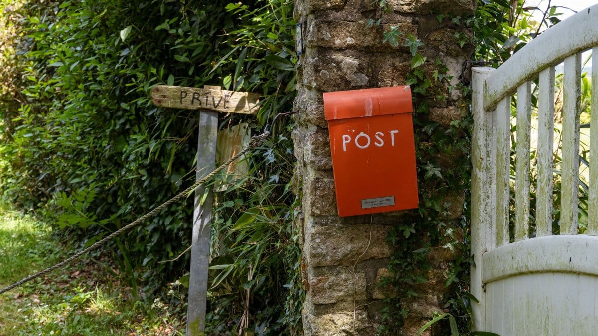 Charming red post box on an ivy-covered stone wall in Brittany, France, near a rustic gate and greenery.
