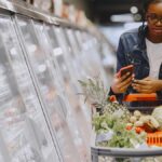 Woman in a grocery store using smartphone while pushing a shopping cart filled with groceries.