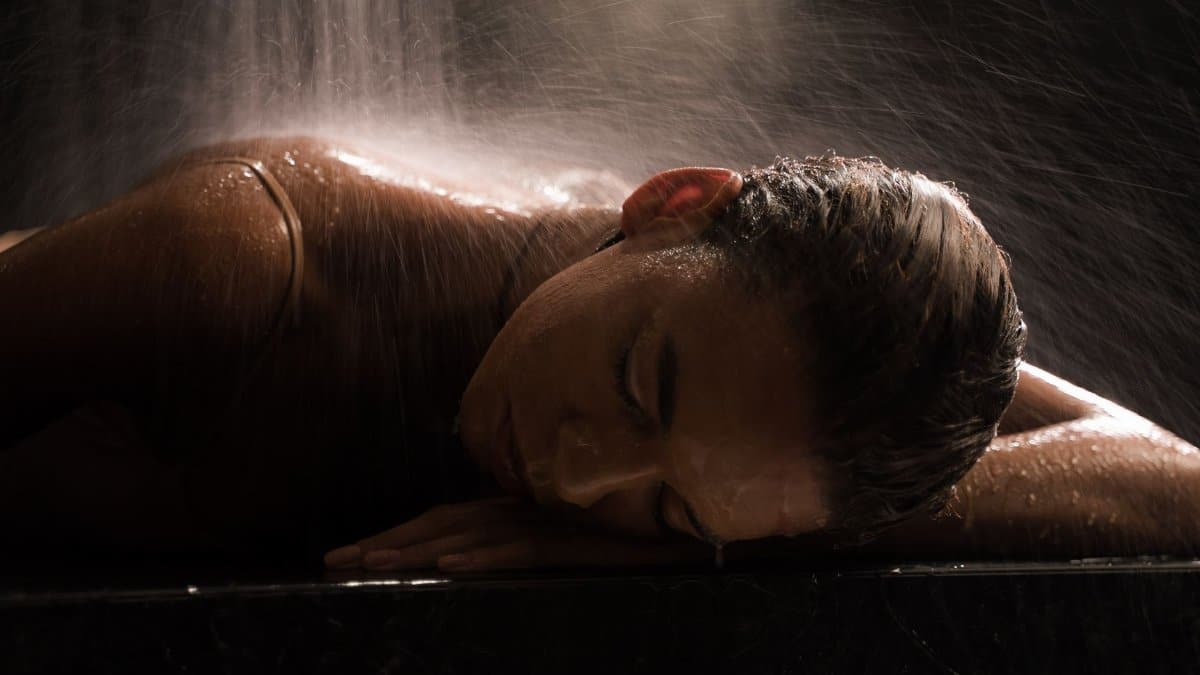 Woman in a spa setting under waterfall, relaxing with eyes closed on a marble surface.