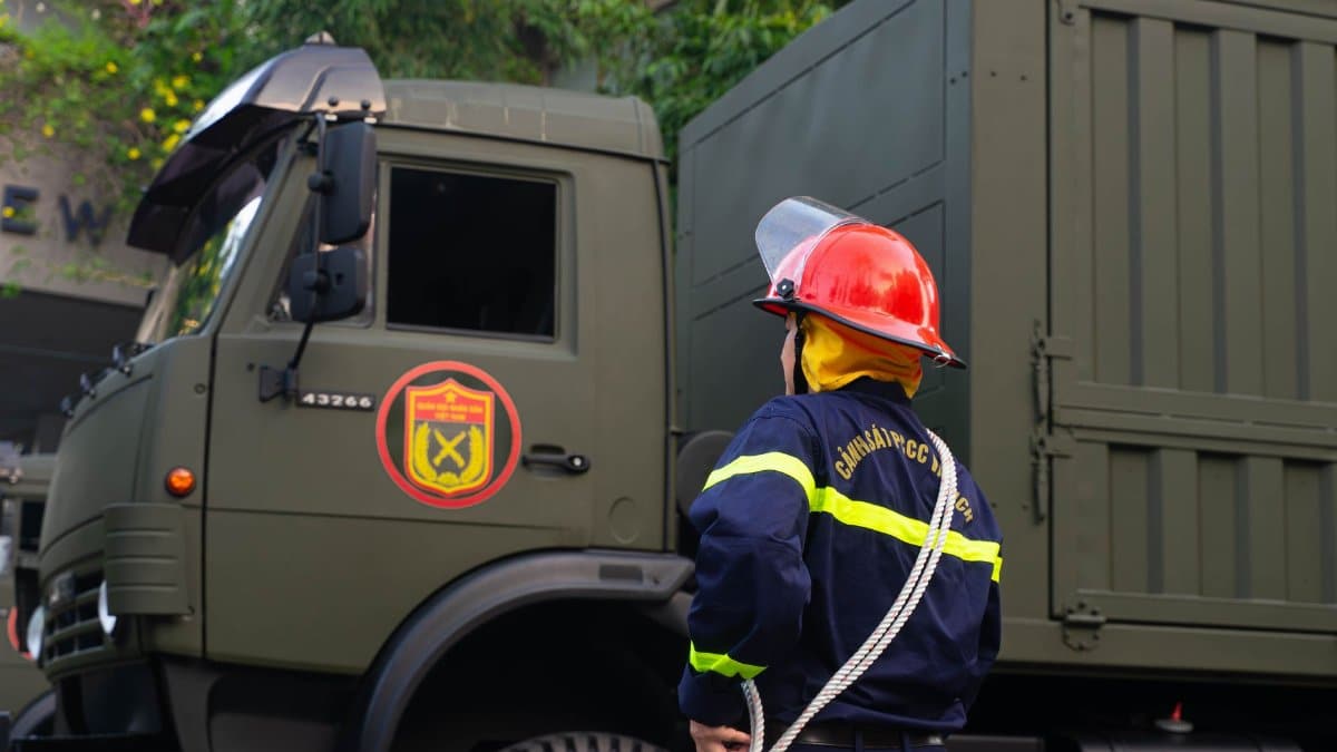 Firefighter in uniform with protective gear stands by an army truck outdoors.