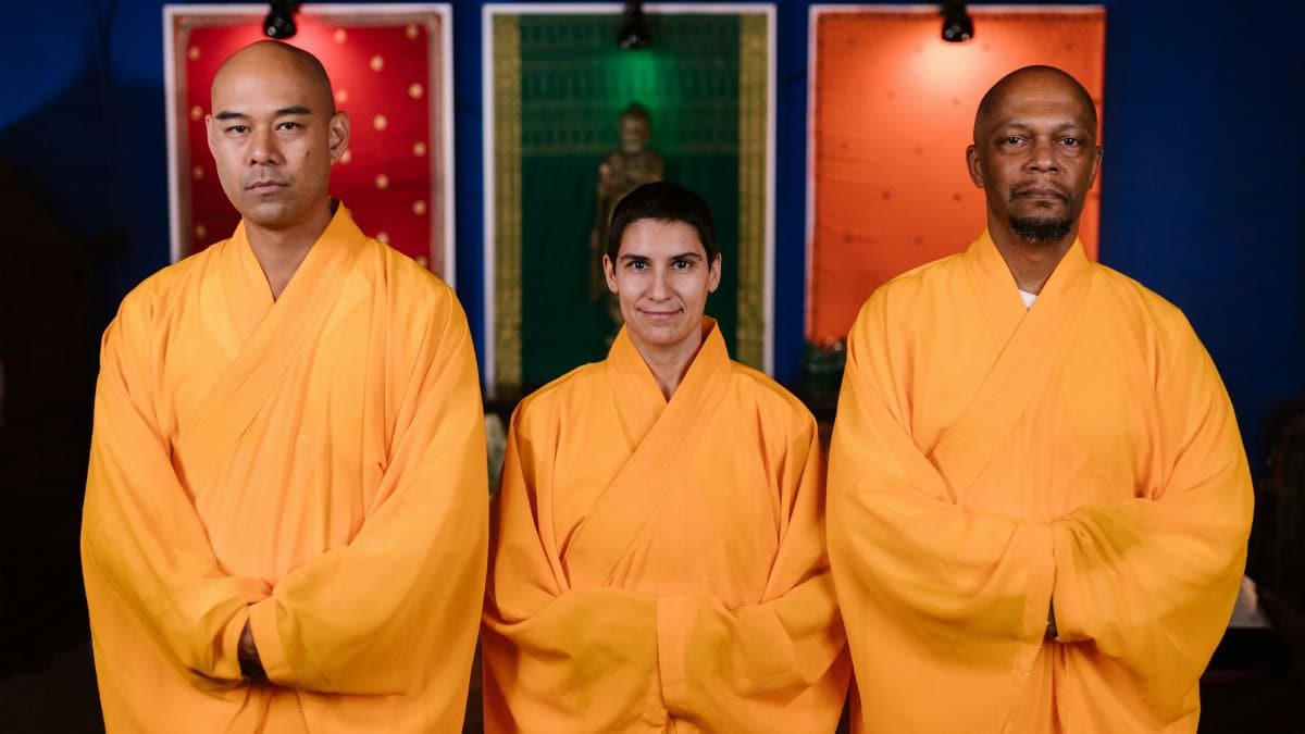 Three Buddhist monks in orange robes pose indoors promoting peace and mindfulness.