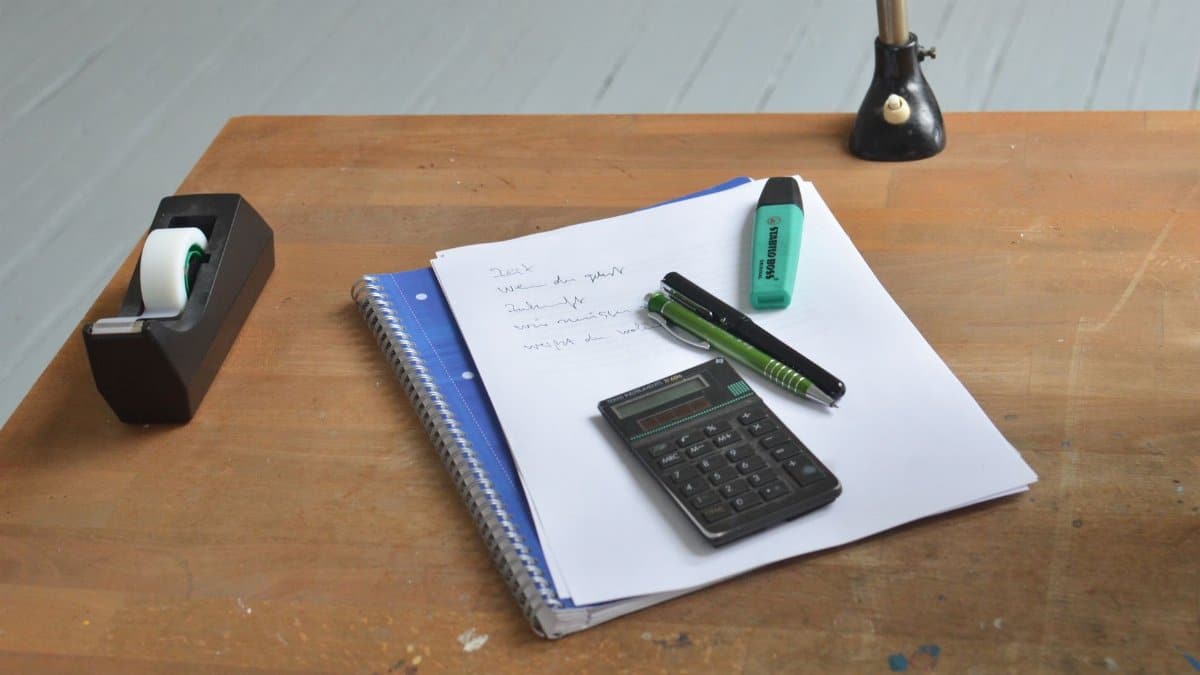 High angle of wooden desk with papers and small calculator with and pens on top near tape dispenser