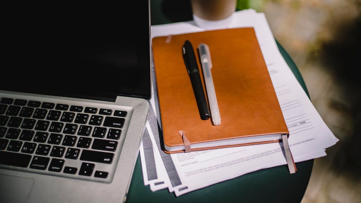 From above of leather notebook with papers and pens placed on table near laptop and cup of coffee
