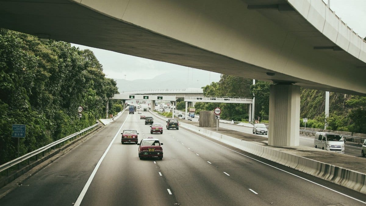 Automobiles driving on asphalt roadway with marking lines under modern bridge in town near trees