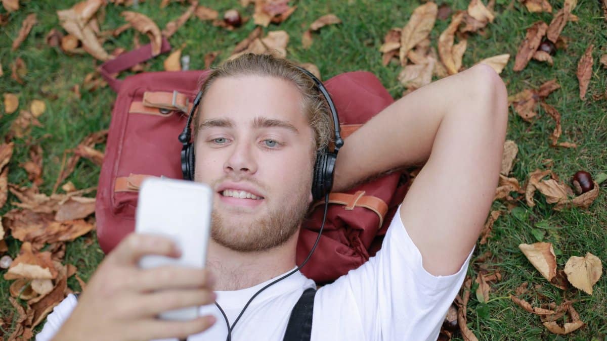 Adult male lying on grass in a park, listening to music with headphones and using a smartphone.