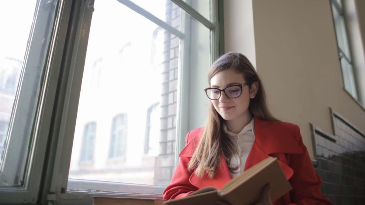 A young woman in a red coat happily reading a book by a window indoors.