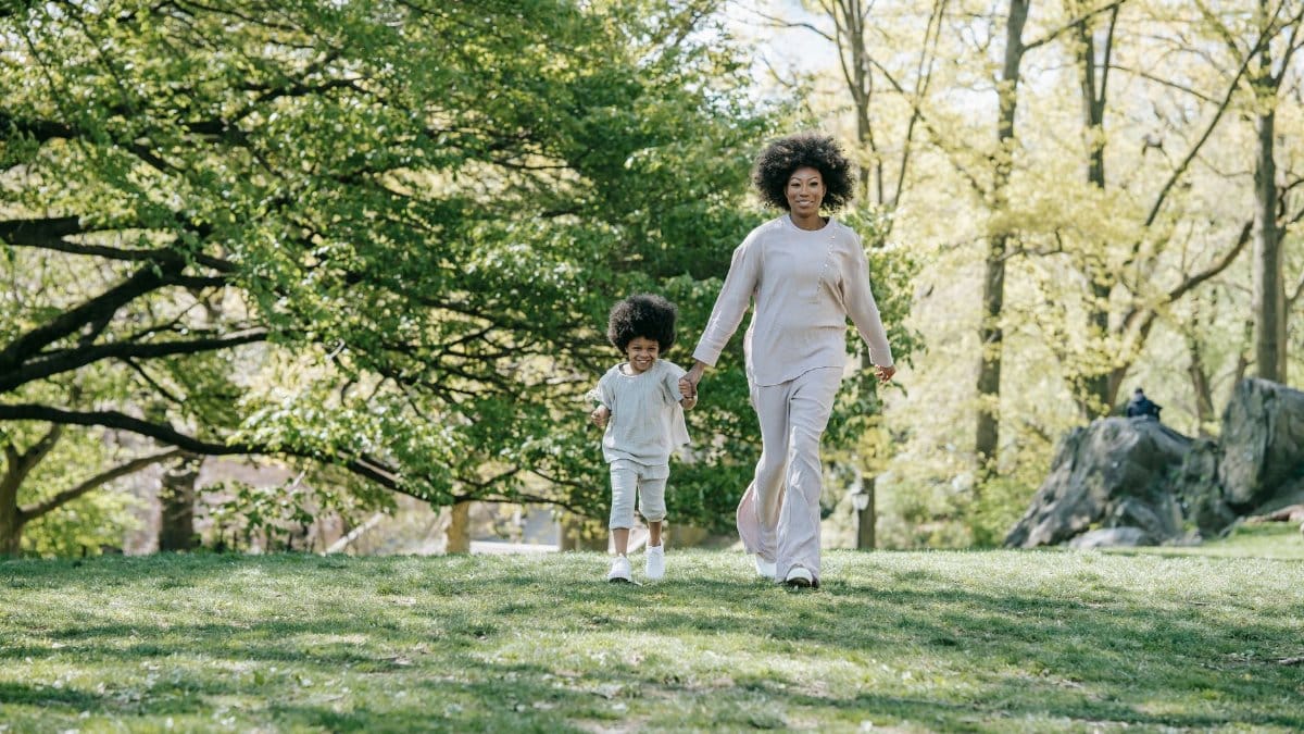 A mother and child holding hands, walking in a sunlit park surrounded by lush greenery.