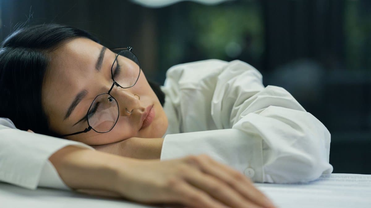 A tired woman with eyeglasses sleeps on a desk indoors, highlighting exhaustion.