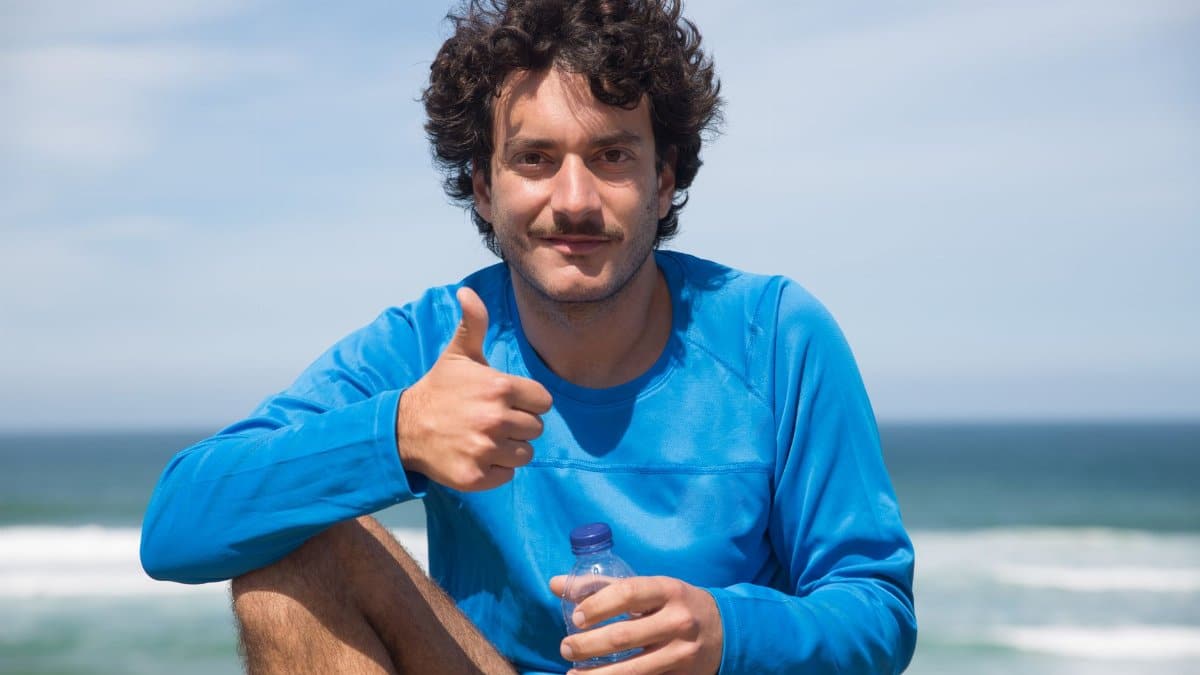 Smiling man in blue long sleeves with curly hair gives a thumbs up at the beach in Portugal.
