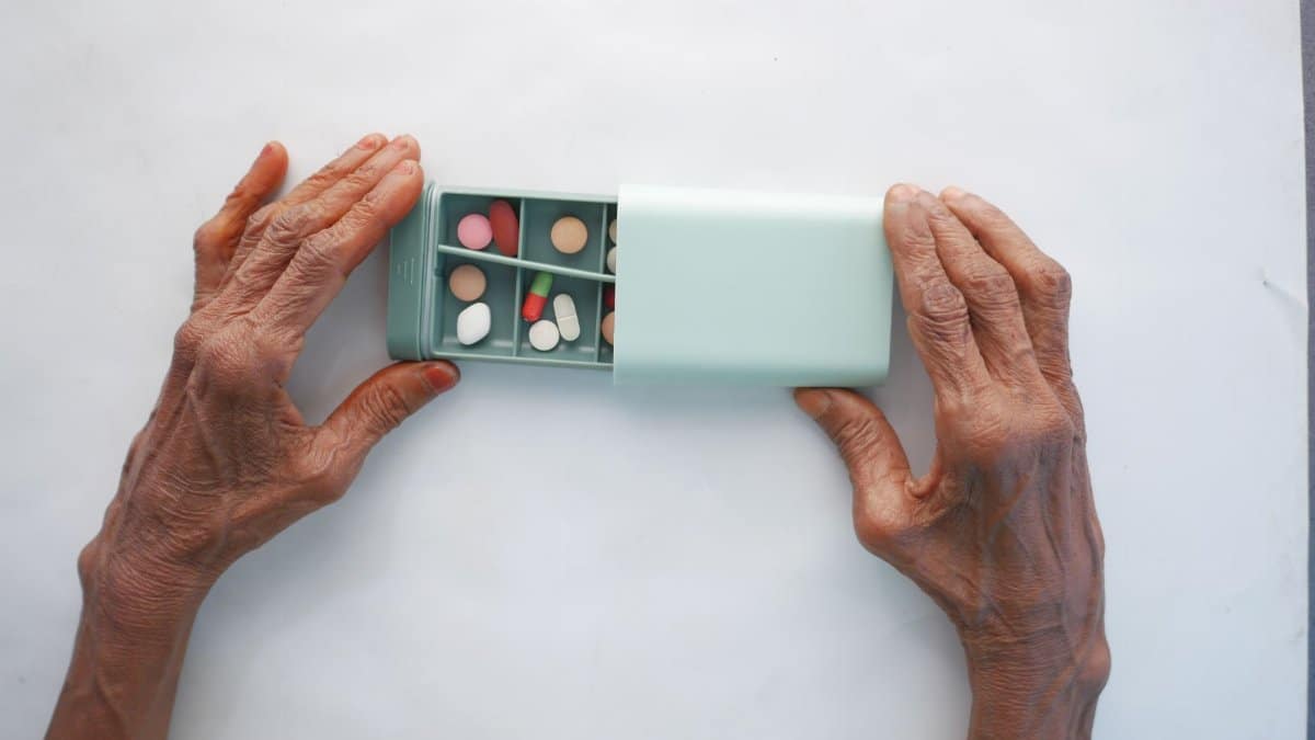 Elderly person sorting pills into a turquoise pillbox on a white surface.
