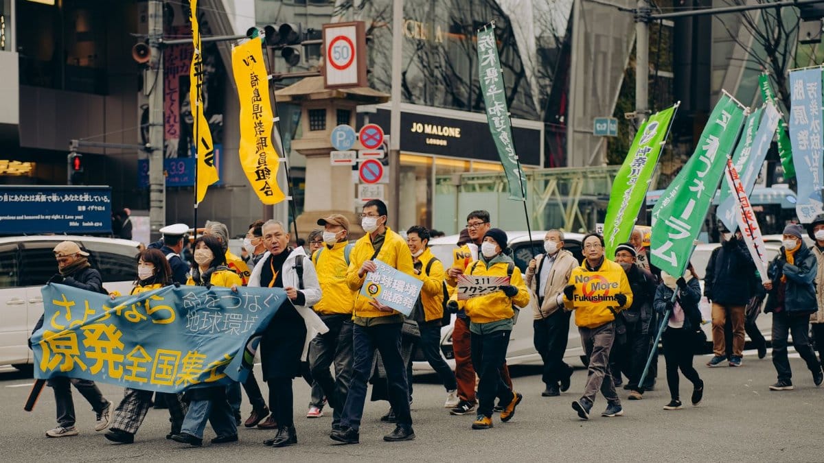 A group of protestors in yellow jackets march through Osaka with colorful flags and banners.