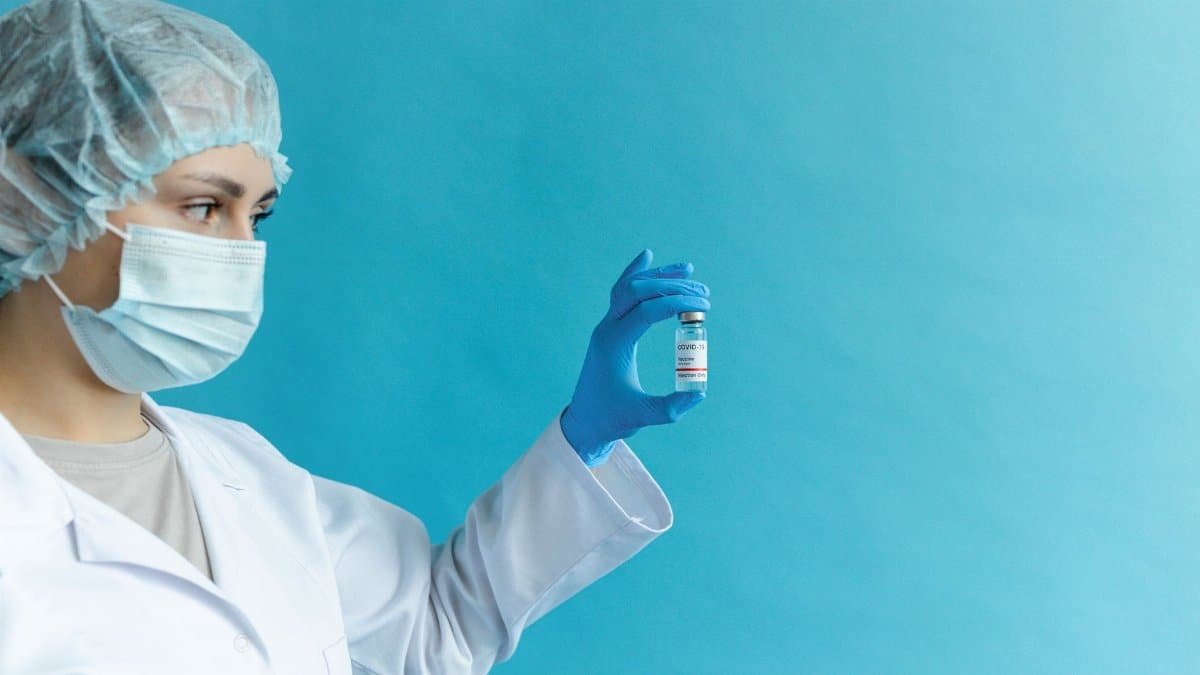 Female doctor in protective gear holding a vaccine vial against a blue background.