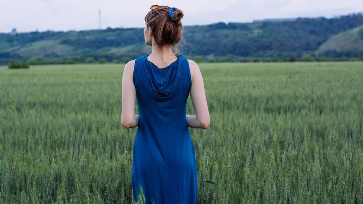 Back view of a woman in a blue dress standing in a vast green grass field.