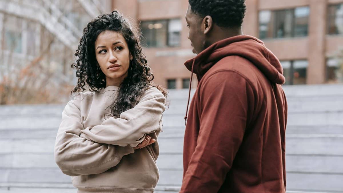 Unset ethnic woman with crossed arms and African American man looking at each other while having quarrel on street with building on blurred background