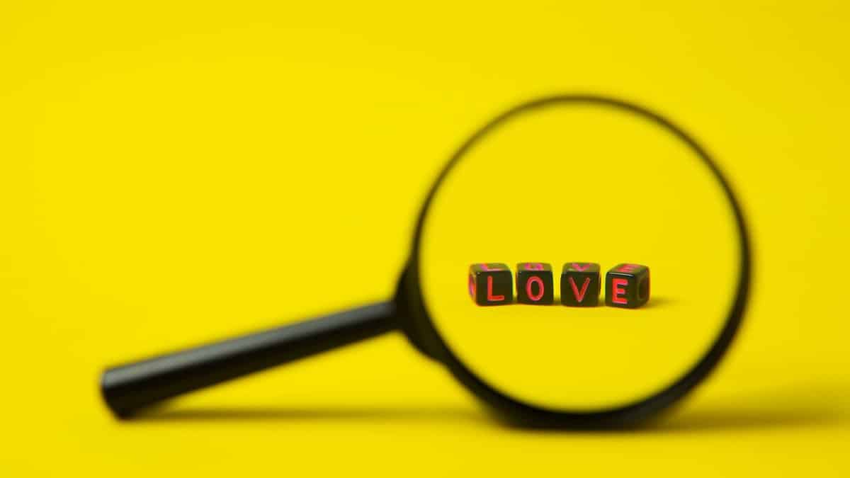 Love sign made of black plastic cubes with letters placed near magnifying glass on bright yellow background