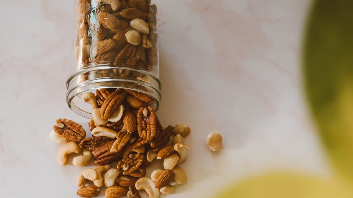 Assorted mixed nuts spill from a glass jar on a light surface, highlighting a variety of textures and colors.