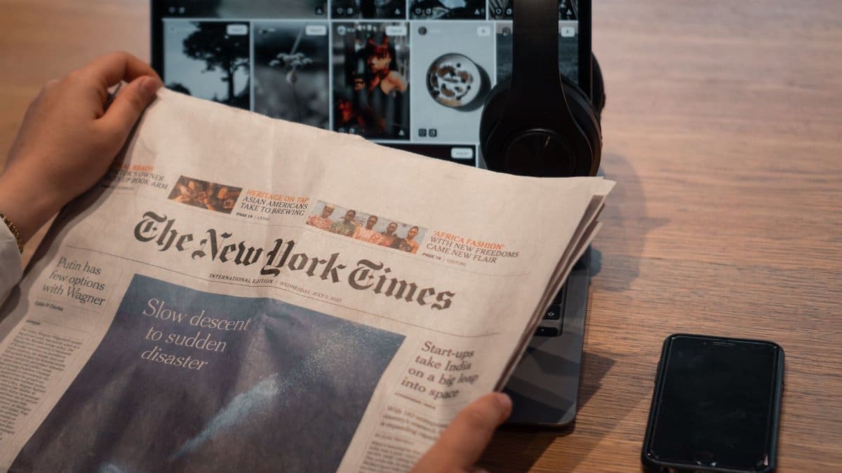 Close-up of hands holding a newspaper by a laptop and smartphone on a table.