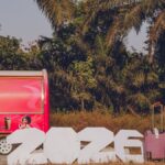 A woman with luggage beside large 2026 sign near a vibrant red trailer outdoors.