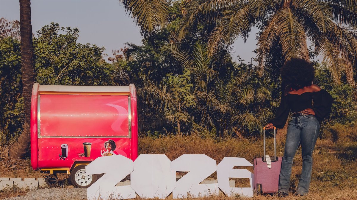 A woman with luggage beside large 2026 sign near a vibrant red trailer outdoors.