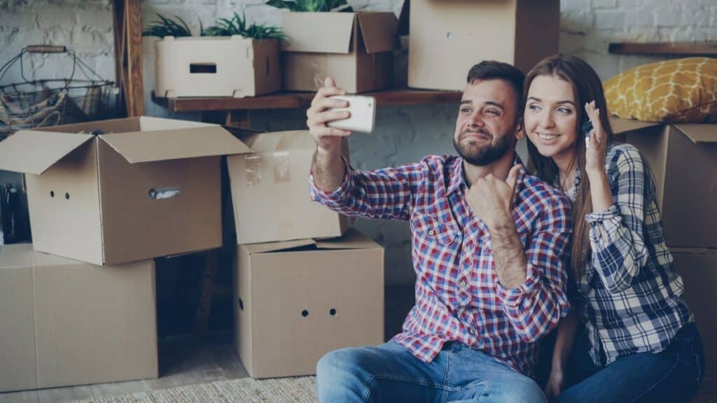 Happy couple taking a selfie while surrounded by moving boxes, celebrating new home.