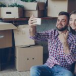 Happy couple taking a selfie while surrounded by moving boxes, celebrating new home.