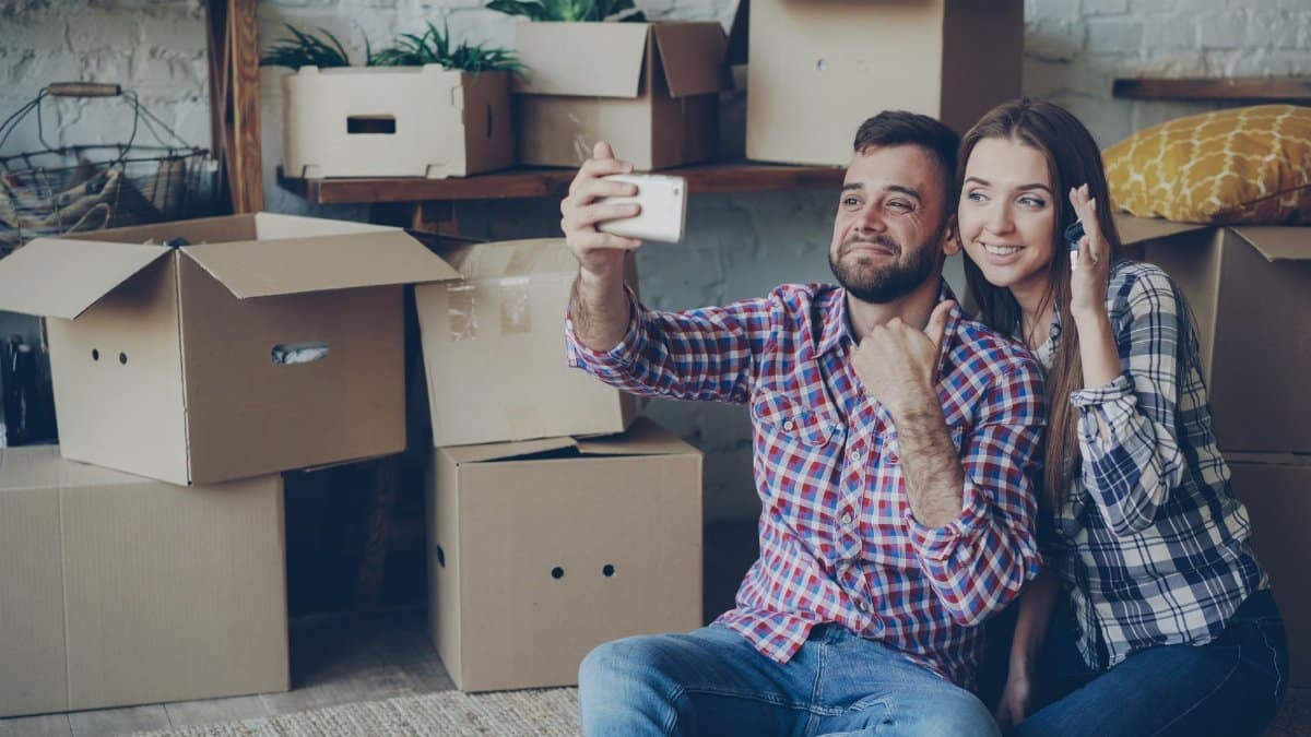 Happy couple taking a selfie while surrounded by moving boxes, celebrating new home.
