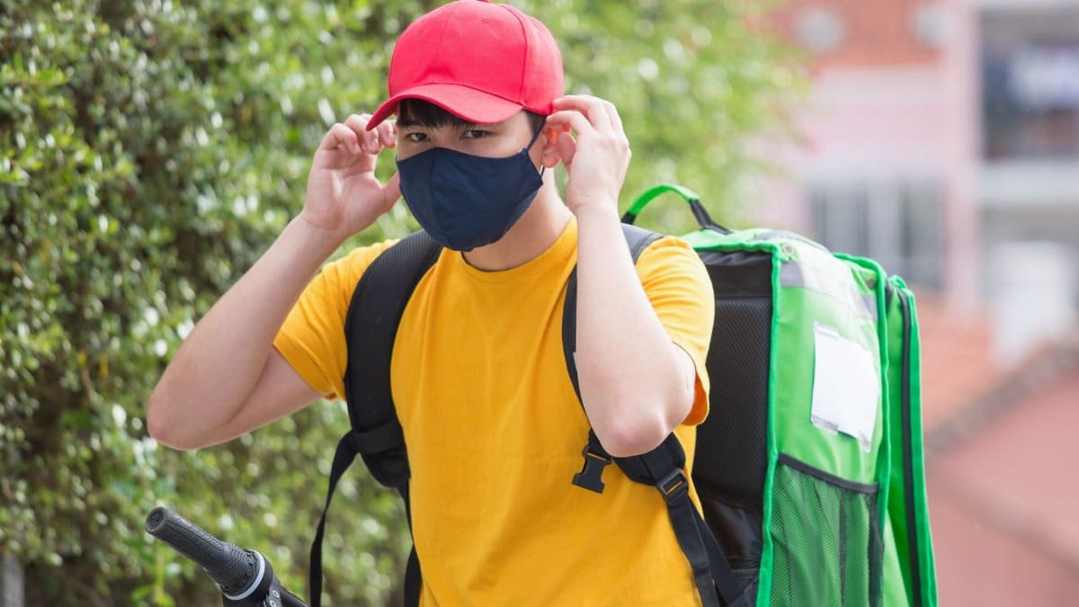 A delivery person adjusts their cap while wearing a face mask and delivering by bike outdoors.