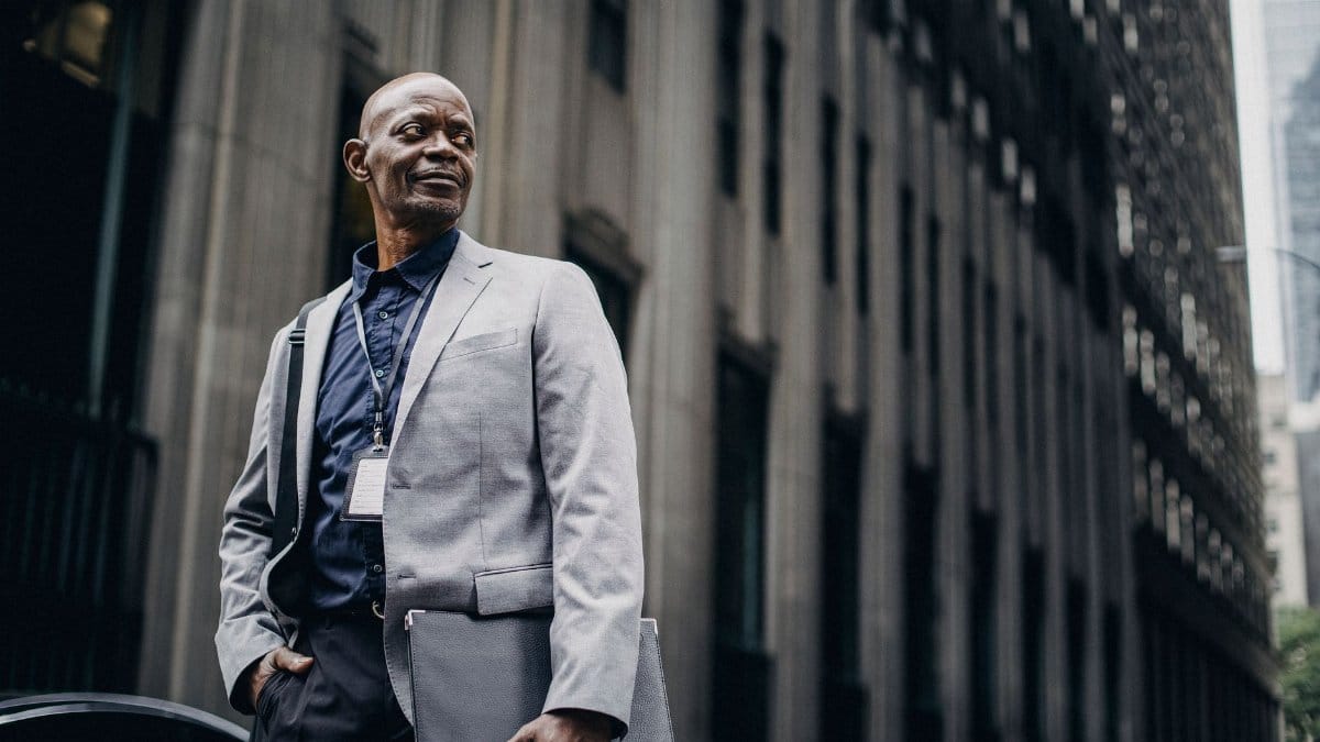 Low angle of confident bald black mature male employee in classy suit and badge standing on city street with folder in hand and looking away