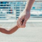 A close-up of a father holding hands with his young daughter outdoors on a sunny day.