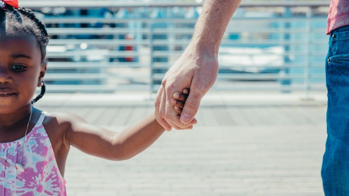 A close-up of a father holding hands with his young daughter outdoors on a sunny day.