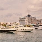 A scenic view of luxury yachts and a cruise ship moored at a bustling port under cloudy skies.