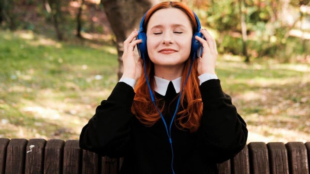 Woman with red hair enjoying music on headphones in a sunny park setting.