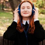 Woman with red hair enjoying music on headphones in a sunny park setting.