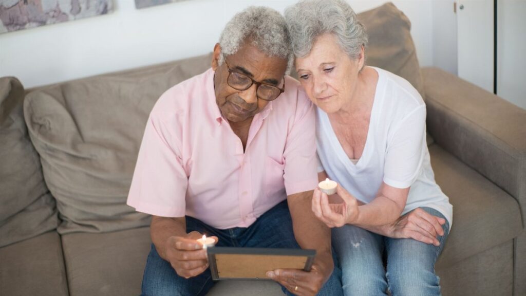 An elderly couple sits on a sofa with a picture frame, reflecting with candles.