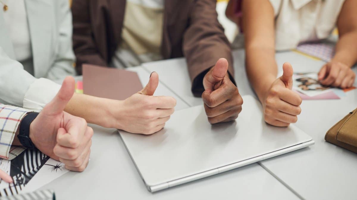 Four diverse hands showing thumbs up over a laptop on a desk, symbolizing teamwork and approval.