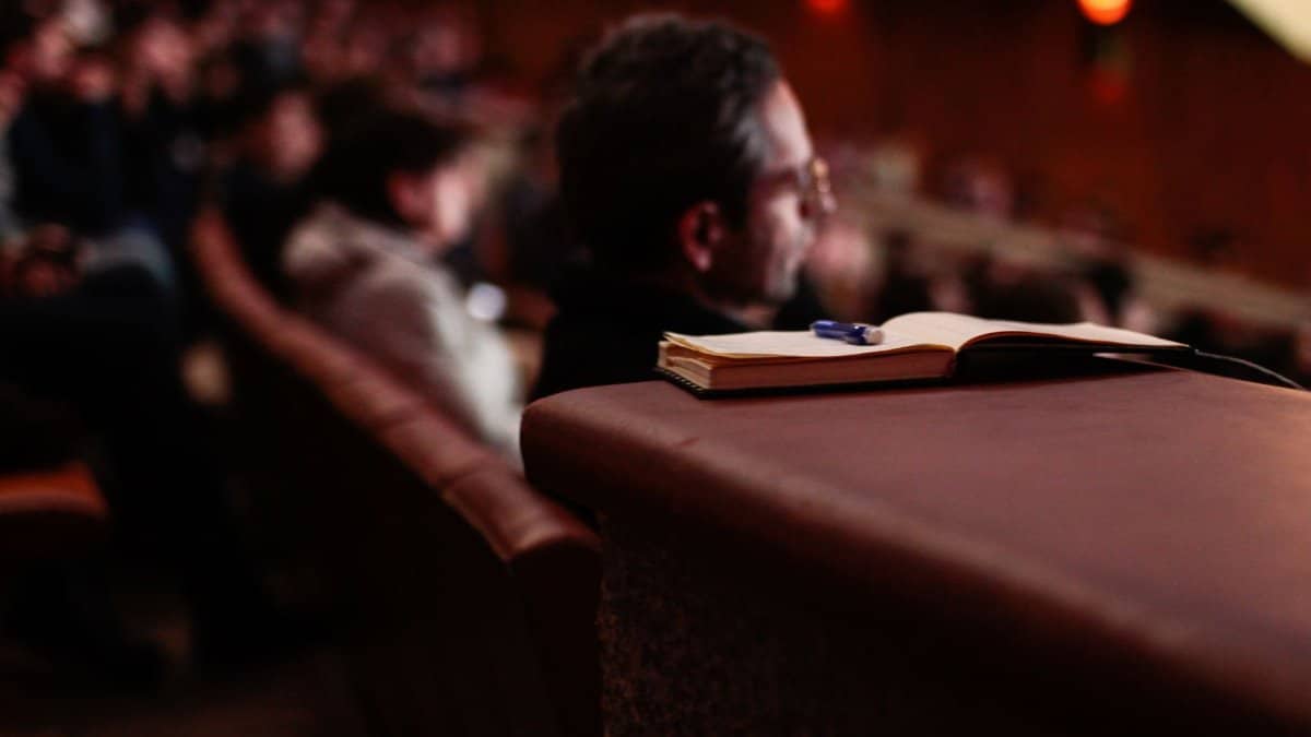 A focused shot of a notebook and pen in an auditorium with blurred audience.