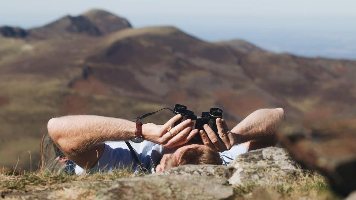 A man lies on a mountainside observing the landscape through binoculars on a clear day.