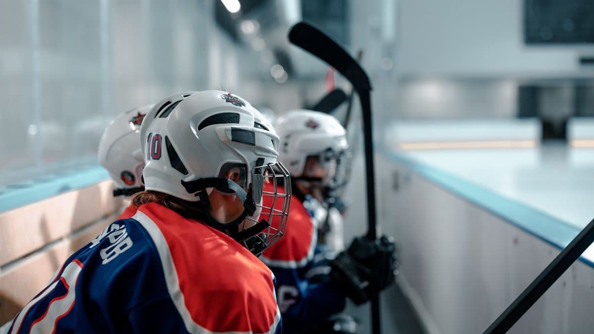 Teen hockey players in helmets and jerseys sit on a rink bench ready to play.