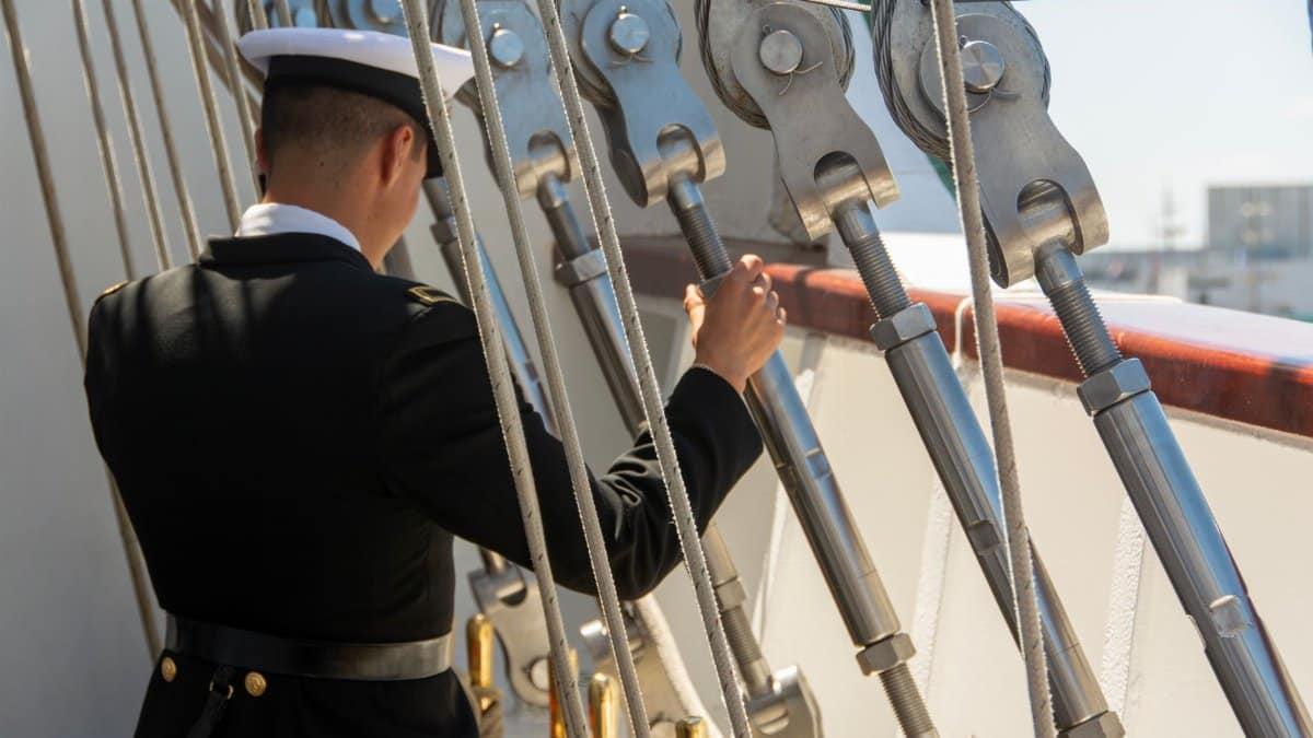 A sailor in uniform adjusts ropes on a ship deck during the day.