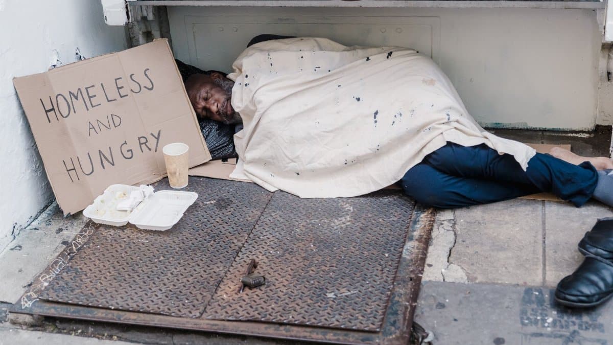 Street scene depicting a homeless man sleeping with a placard reading 'Homeless and Hungry'.