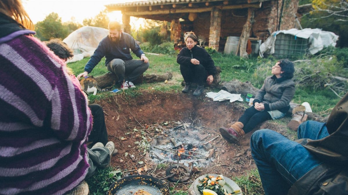 A group of adults seated around a campfire near a rustic farmhouse enjoying a meal.