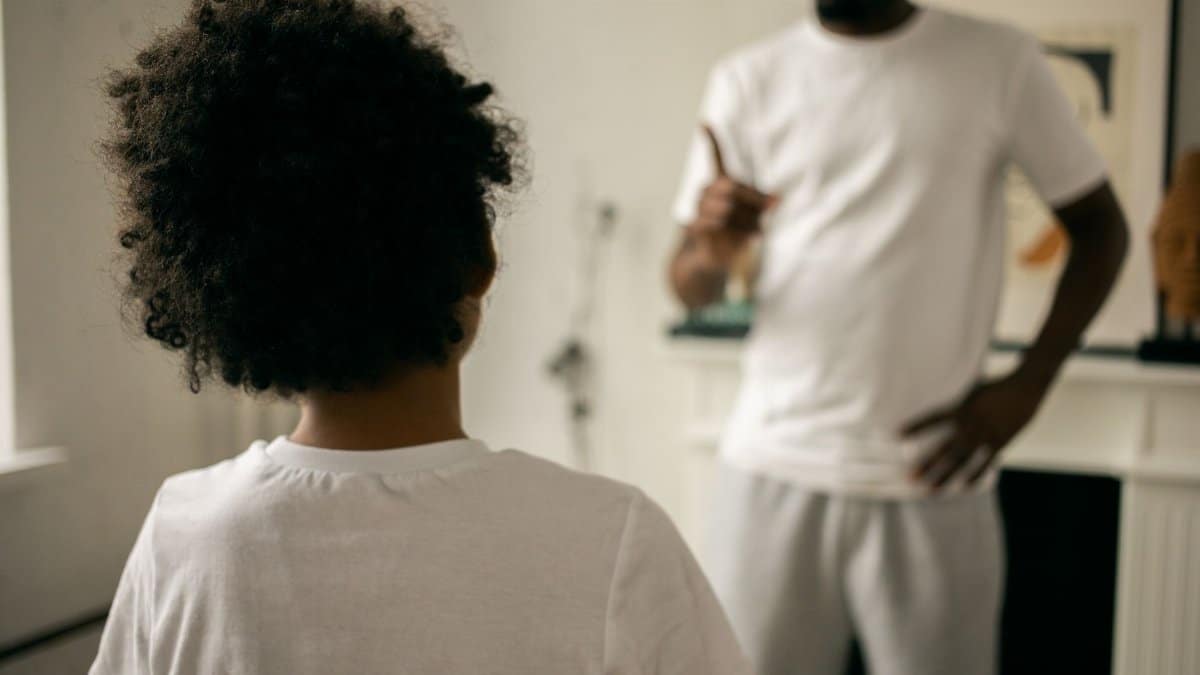 Back view of unrecognizable black boy with Afro hair standing in front of crop anonymous dad pointing finger and scolding at home