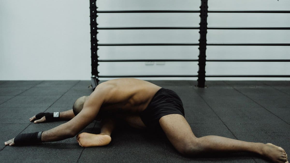 Shirtless man stretches while sitting on gym floor, showcasing flexibility.